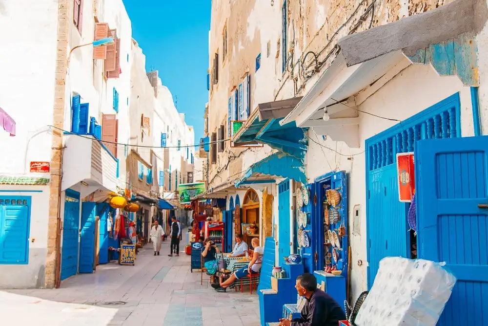 A colorful street in the historic medina of Essaouira, Morocco, lined with traditional shops, vibrant blue doors, and locals enjoying the sunny day.